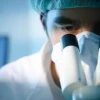 Researcher looks into a microscope wearing mask, hairnet and lab coat.