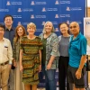 Denise Roe poses for a photo with six of her colleagues in front of a U of A Cancer Center blue backdrop next to a sign announcing her retirement.