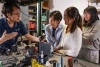 A professor with dark, straight short hair and glasses shows a small black device to three students who are watching the speaker intently.  