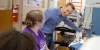 Two people in a lab look at each other and smile. One is standing with a blue button down shirt and short hair, and one person has long brown hair in a clip and is wearing a purple T-shirt