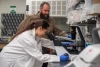 A person in a lab coat with long brown hair in a ponytail  and blue latex gloves looks into a microscope. Another person with a beard and brown button down shirt watches from the side.